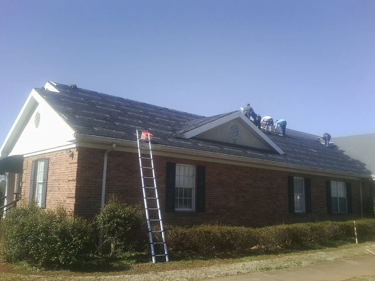 Crew tearing shingles off a brick ranch roof with a ladder set — Metal Roof Repair work by our crew in Lake Odessa