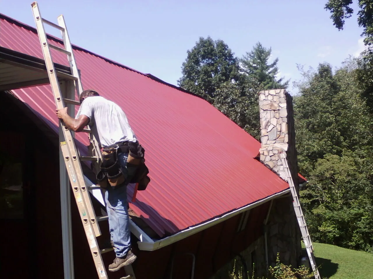 Roofer on a ladder beside a red metal roof and stone chimney — Soffit & Fascia Repair by Roofing Brothers in Lake Odessa
